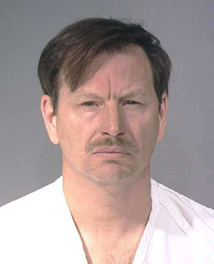A white man with brown hair and a mustache looks directly at the camera wearing white prison clothes.