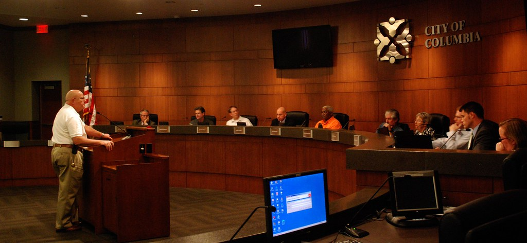Columbia, Missouri Police Chief speaks to the Citizens Police Review Board. The Chief stands at a podium facing ten men and women Board members seated behind a counter.